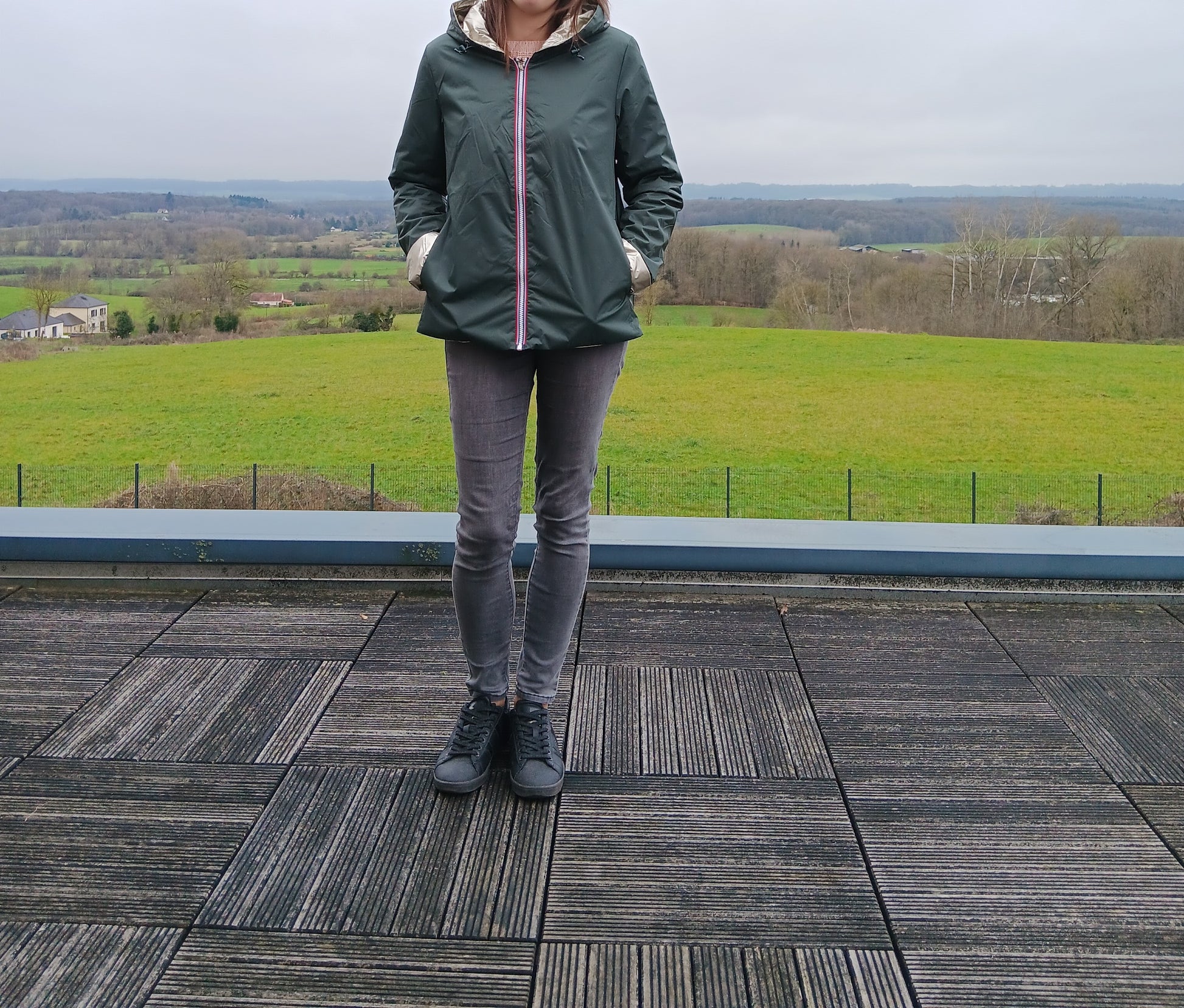 Imperméable réversible vert forêt avec garniture en ovale doré sur le torse, porté par une personne debout sur une terrasse extérieure avec paysage champêtre en arrière-plan.