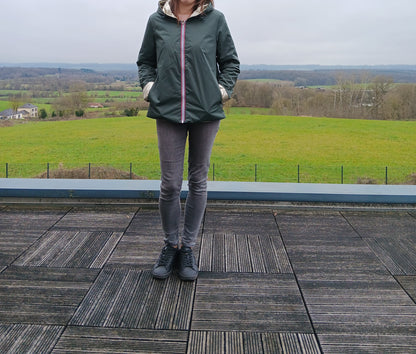 Imperméable réversible vert forêt avec garniture en ovale doré sur le torse, porté par une personne debout sur une terrasse extérieure avec paysage champêtre en arrière-plan.