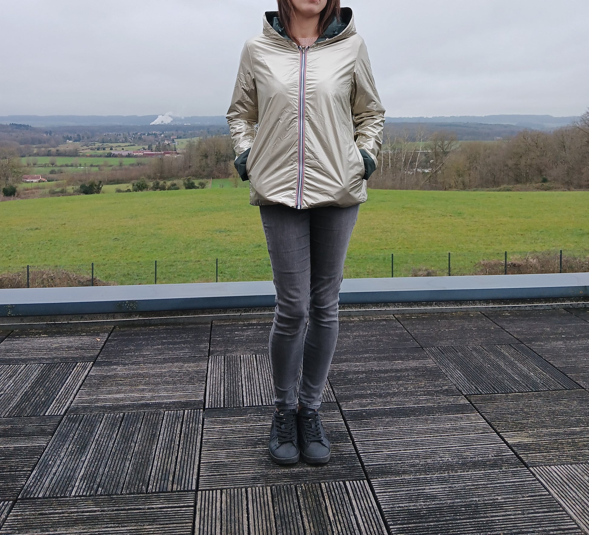 Imperméable réversible court couleur beige doré avec capuche, garnissage ouate, porté par une personne debout sur une terrasse, paysage rural en arrière-plan.