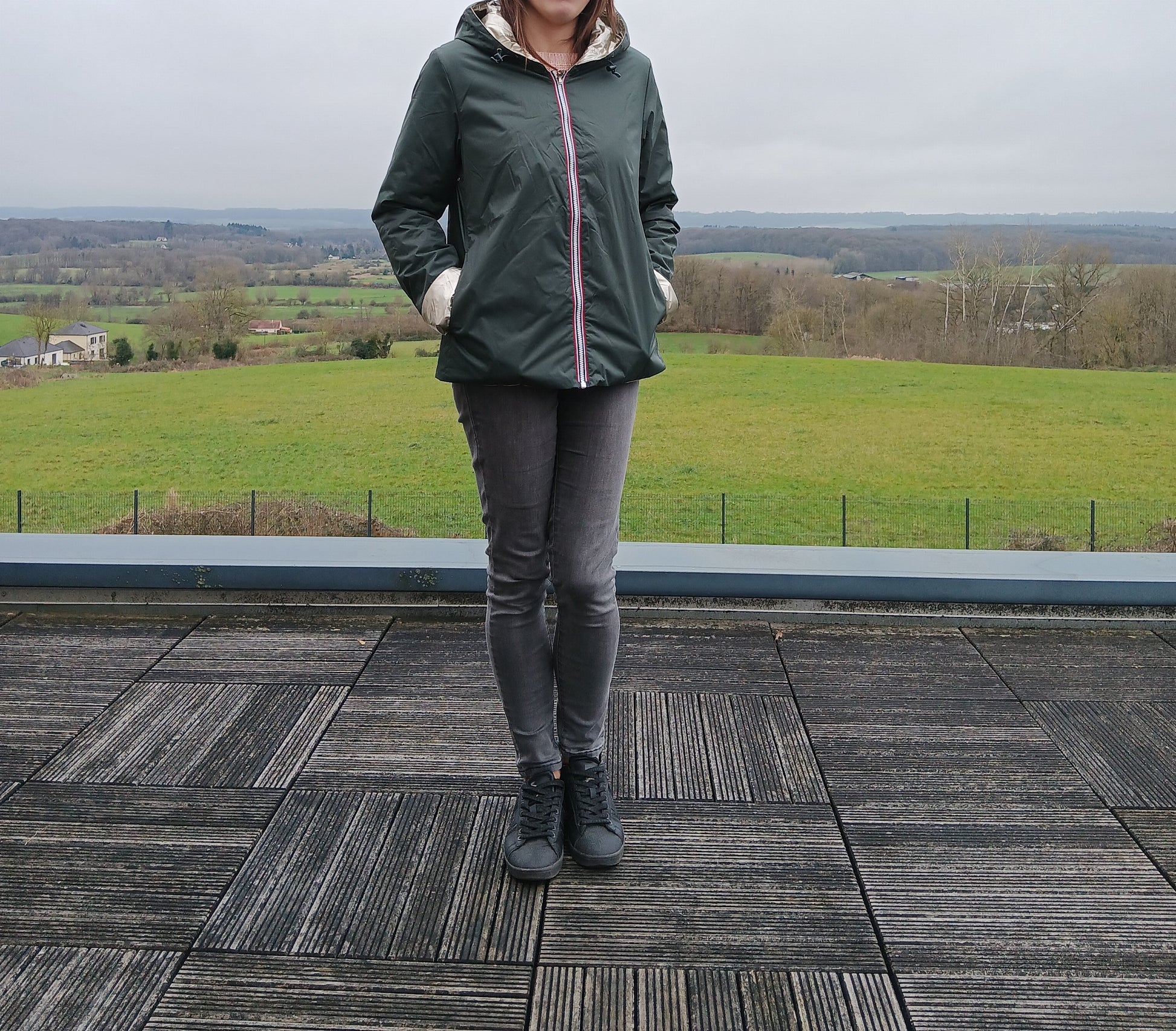 Laura Jo – imperméable réversible court vert forêt avec intérieur doré et garnissage en ouate, vue de femme debout sur terrasse ouverte au paysage rural.