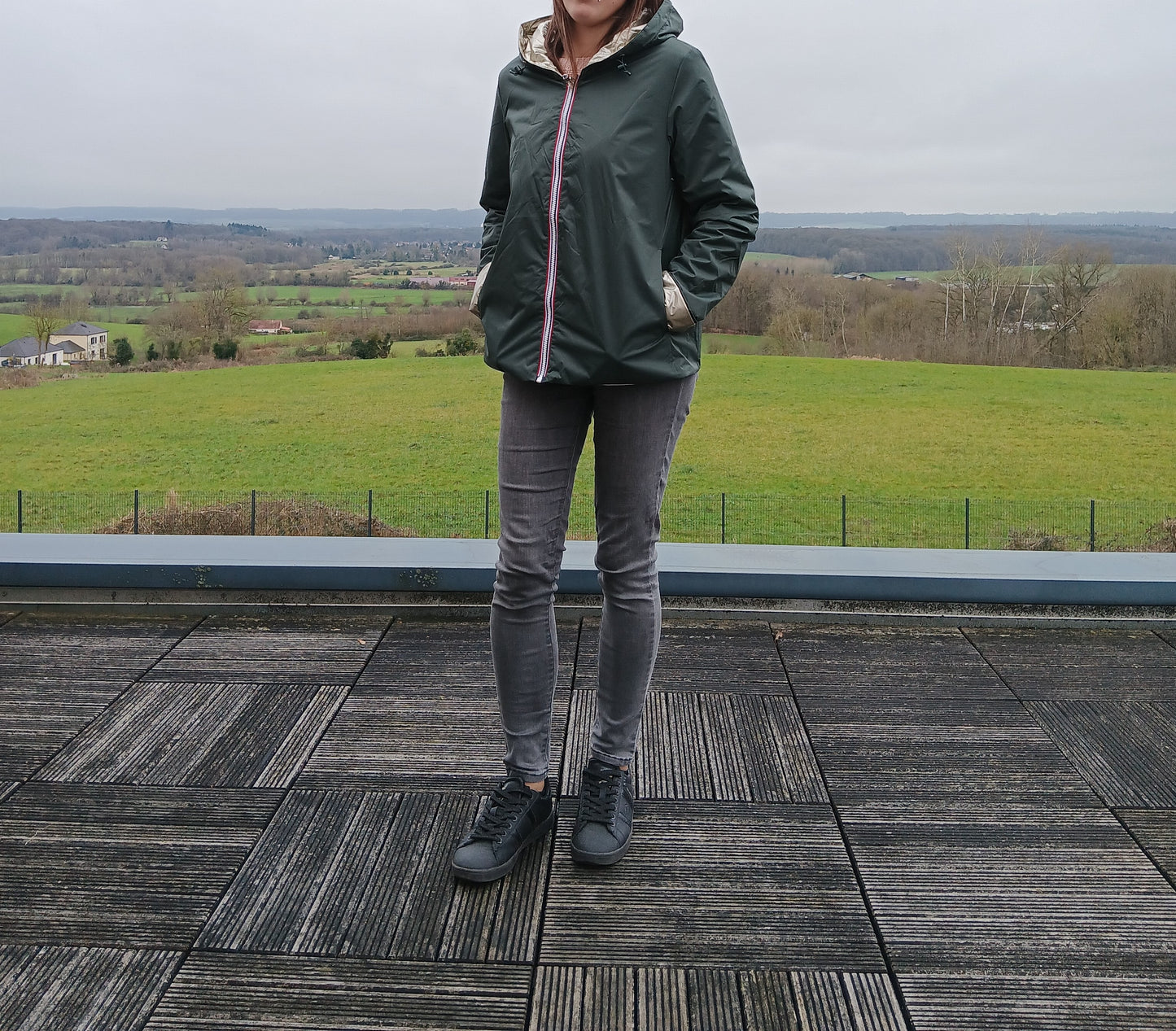 LaurA Jo imperméable réversible court vert forestier avec garniture en ouate, porté avec jeans gris et sneakers sur terrasse avec paysage rural.