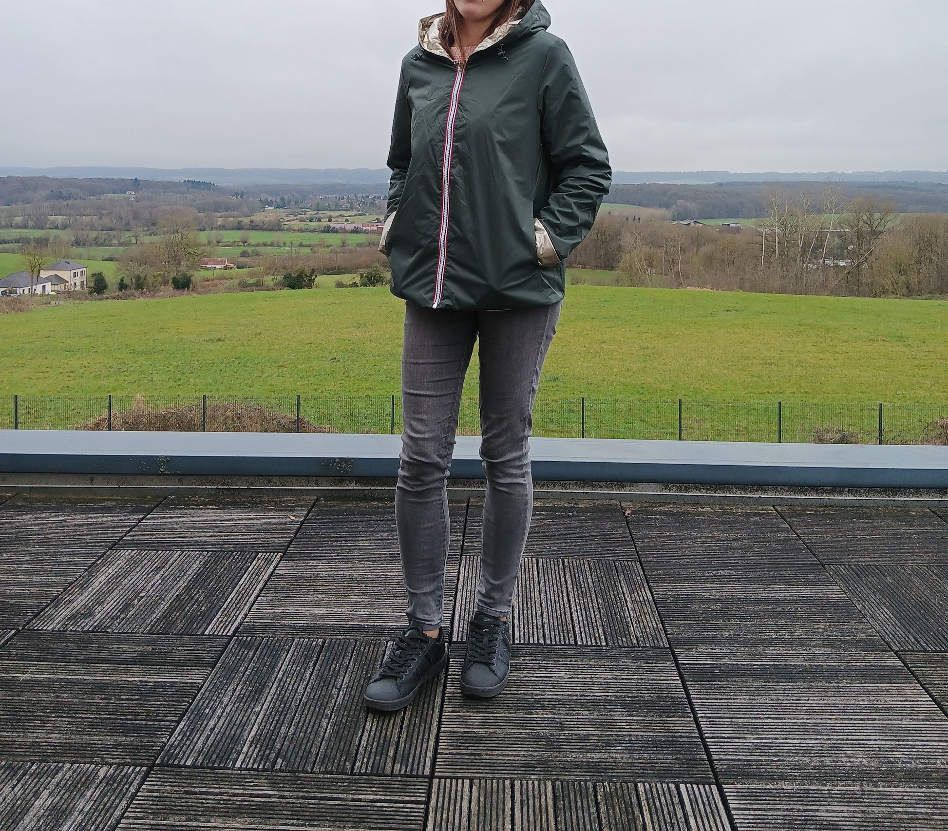 LaurA Jo imperméable réversible court vert forestier avec garniture en ouate, porté avec jeans gris et sneakers sur terrasse avec paysage rural.