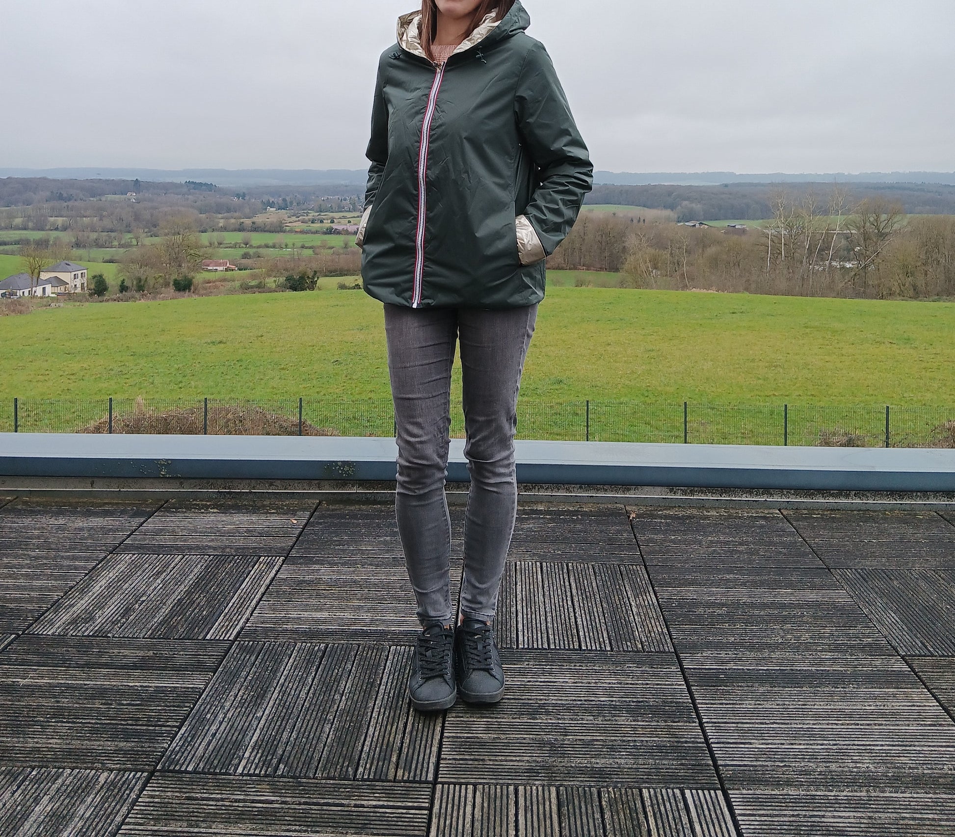 Imperméable réversible vert forêt avec garniture en ouate, person standing outdoors sur terrasse, avec paysage rural en arrière-plan.