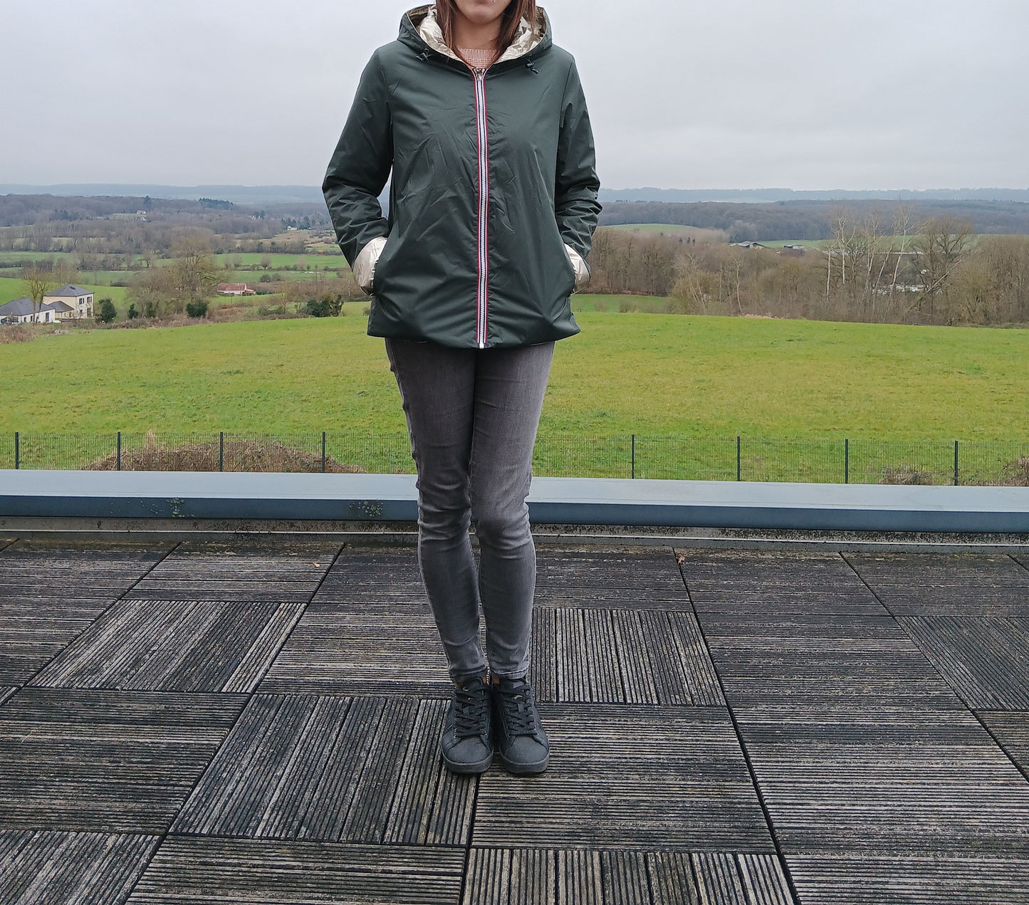 Homme ou femme debout sur une terrasse en bois, portant un imperméable réversible vert forêt avec garnissage en ouate, capuche et poches.
