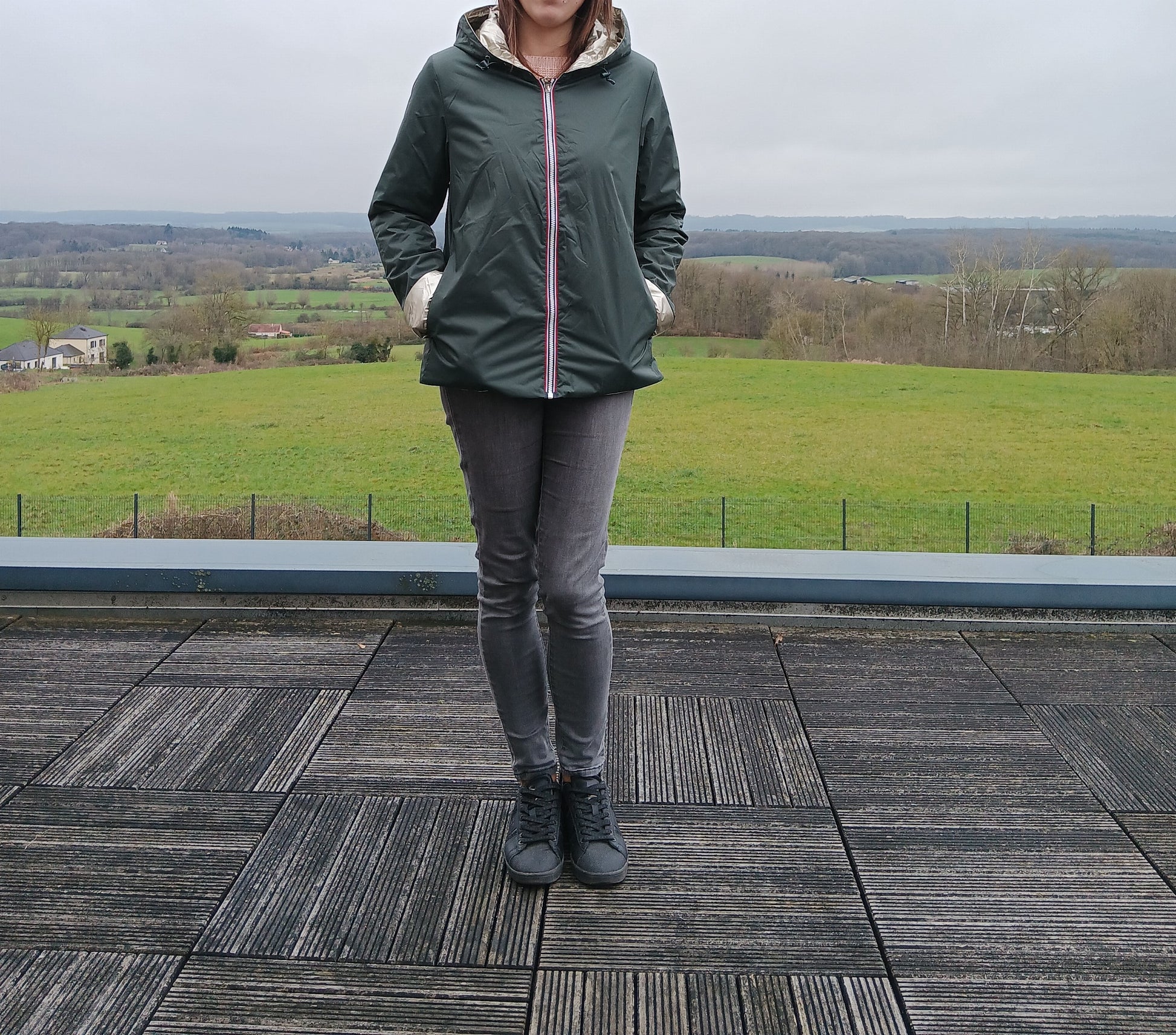 Homme ou femme debout sur une terrasse en bois, portant un imperméable réversible vert forêt avec garnissage en ouate, capuche et poches.