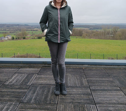 Homme ou femme debout sur une terrasse en bois, portant un imperméable réversible vert forêt avec garnissage en ouate, capuche et poches.