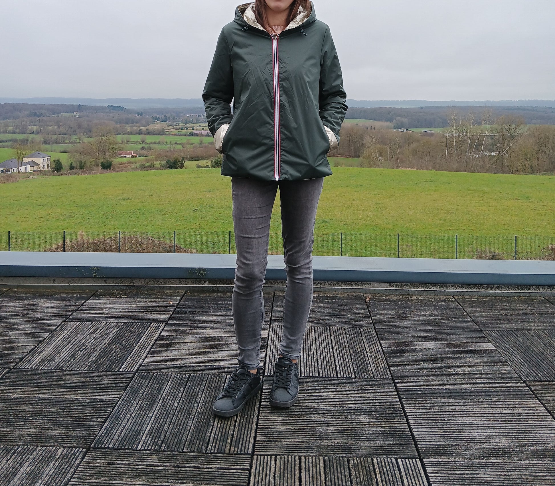 Laura Jo: imperméable réversible court en vert forêt avec garnissage ouate, fermeture zippée, manches et capuche visibles, porté en extérieur sur fond paysage rural.