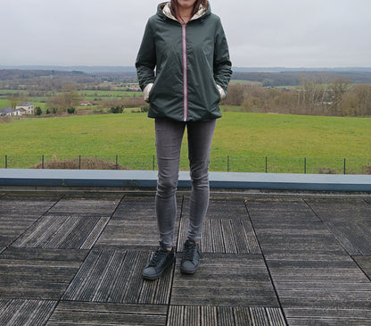 Laura Jo: imperméable réversible court en vert forêt avec garnissage ouate, fermeture zippée, manches et capuche visibles, porté en extérieur sur fond paysage rural.