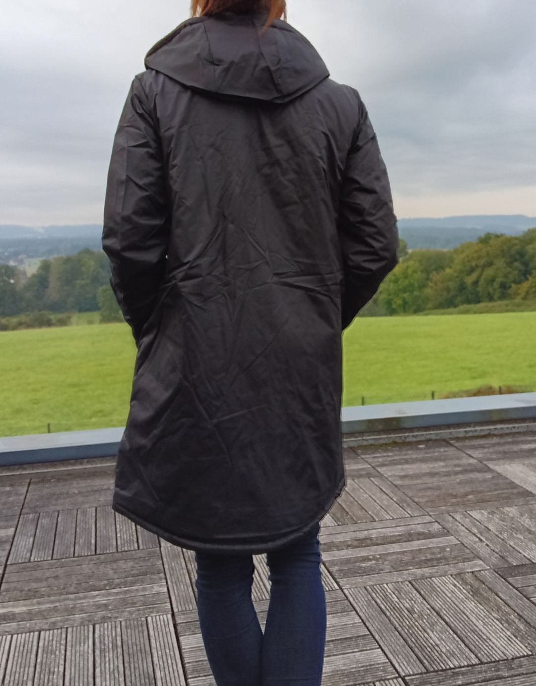 Woman wearing black Laura Jo long raincoat on wooden terrace with green field view