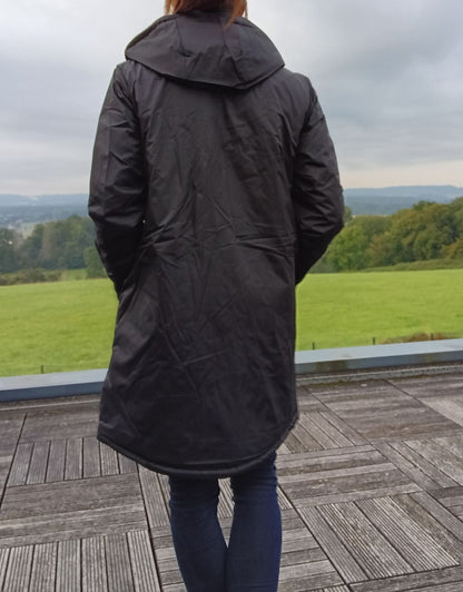 Woman wearing black Laura Jo long raincoat on wooden terrace with green field view