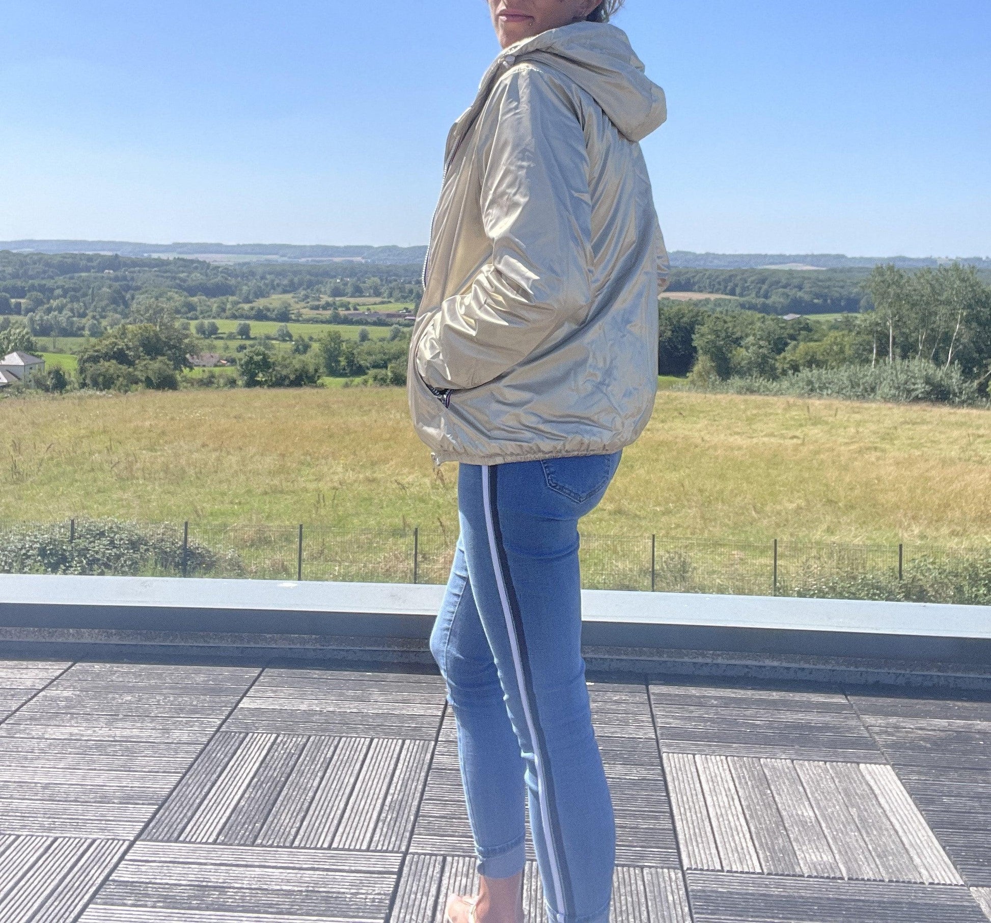 Woman in Laura Jo short waterproof gold jacket and jeans on a terrace, countryside view