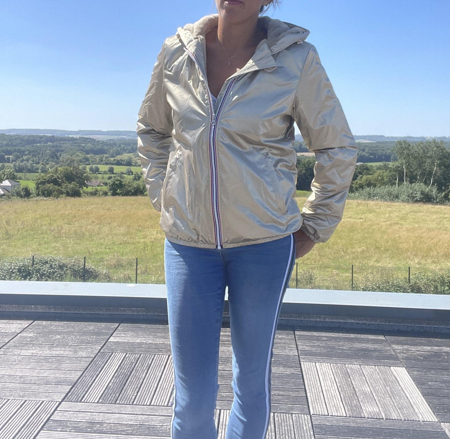 Woman wearing Laura Jo gold waterproof short jacket on terrace with scenic countryside view