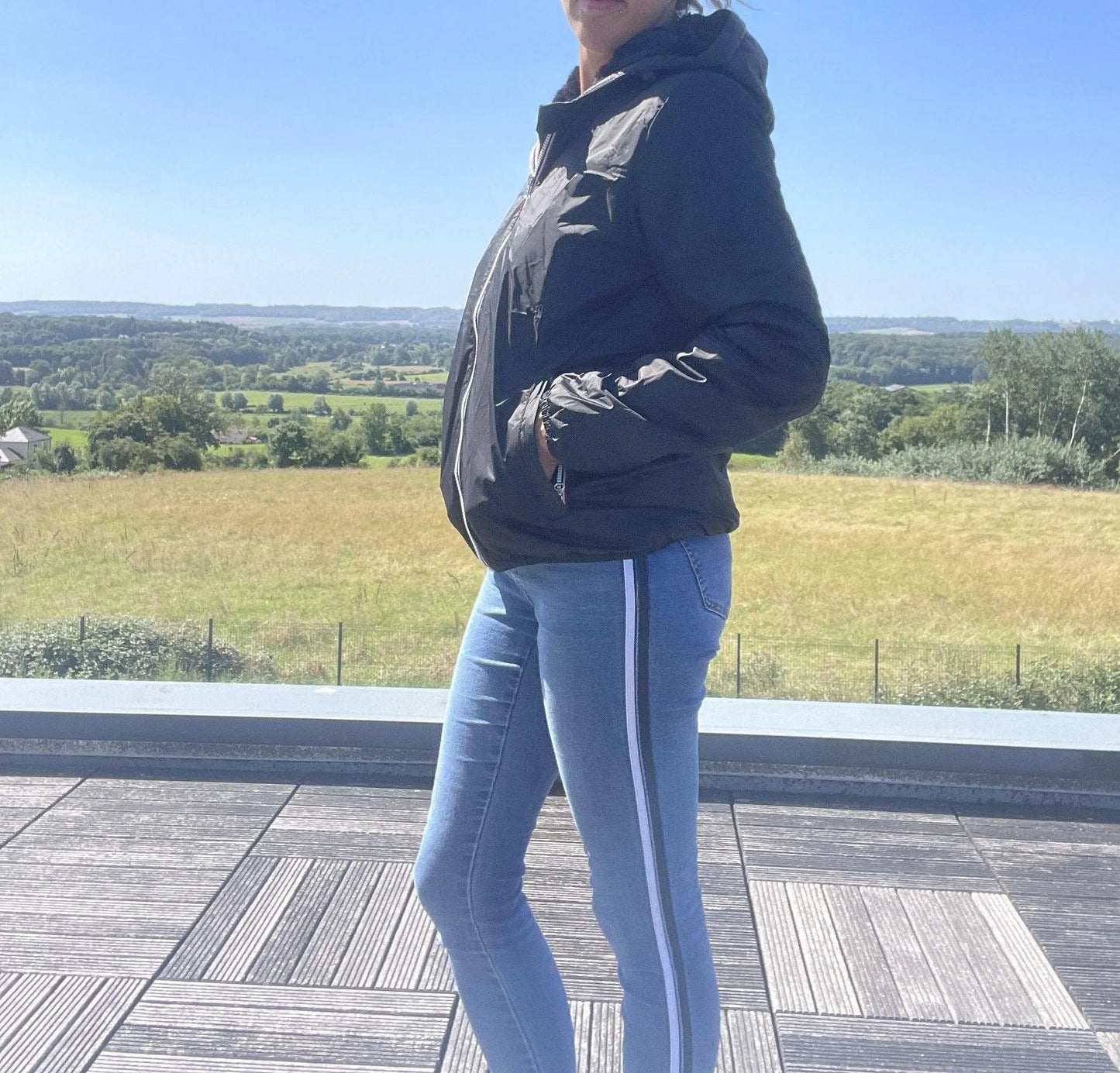 Woman wearing black Laura Jo raincoat and blue jeans on a terrace with scenic countryside