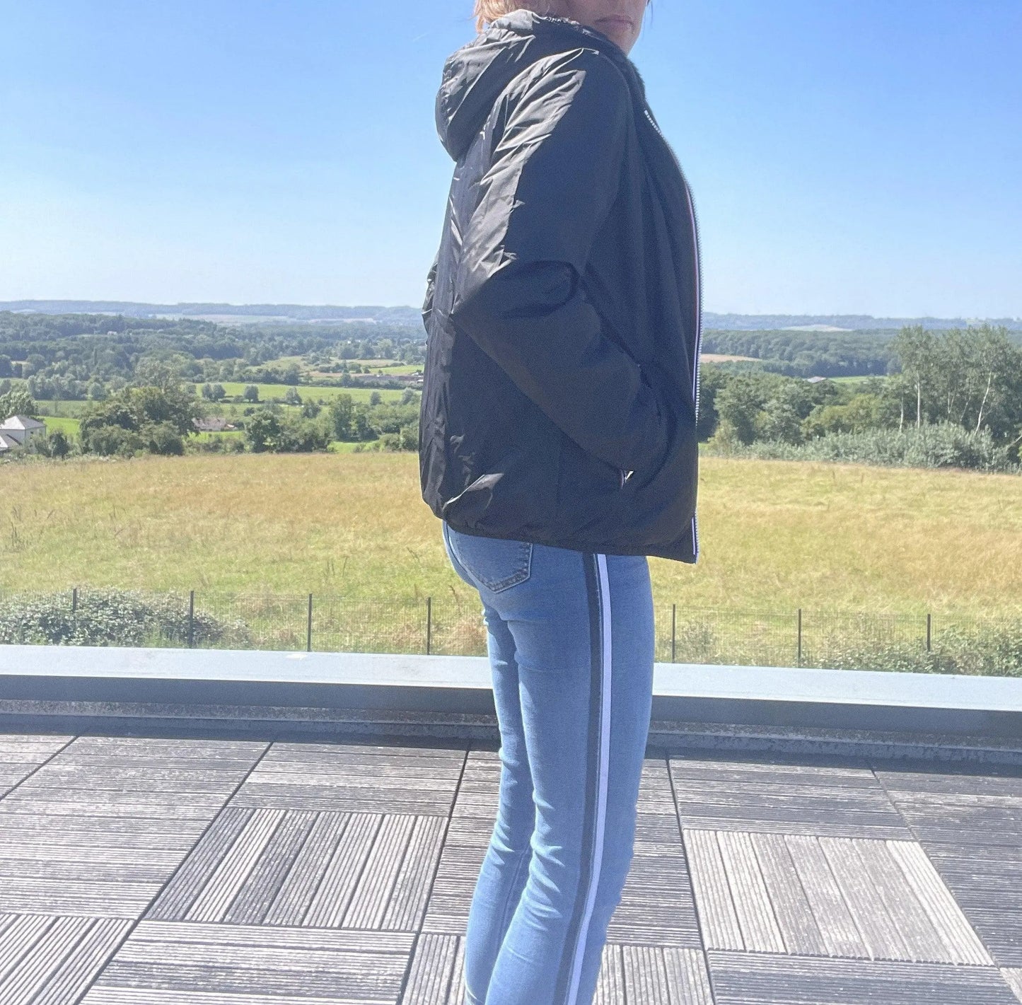 Woman in black Laura Jo raincoat and blue jeans on terrace, countryside background, Le Petit Placard