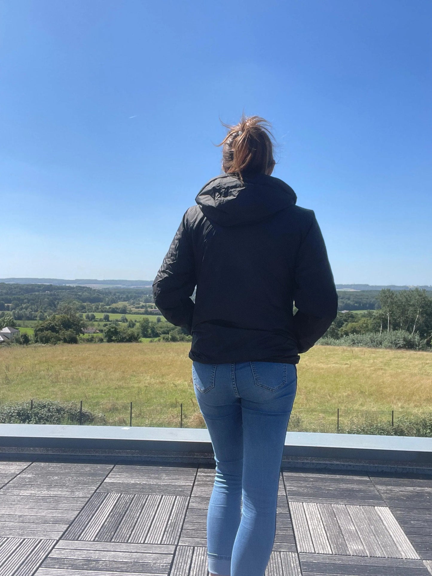 Woman in Laura Jo rain jacket and jeans on terrace overlooking countryside under blue sky