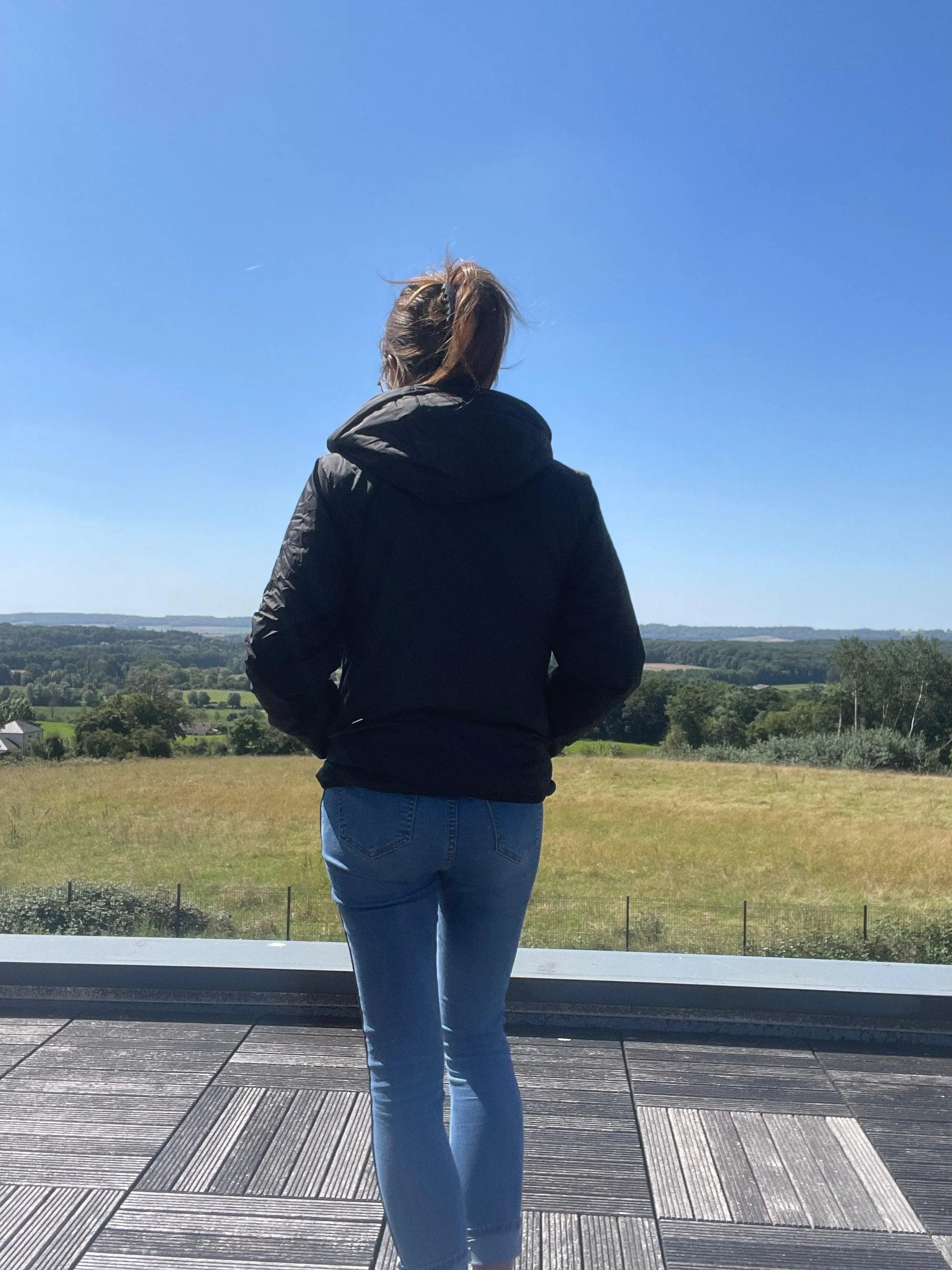 Woman in Laura Jo raincoat and jeans on terrace, overlooking green landscape under blue sky