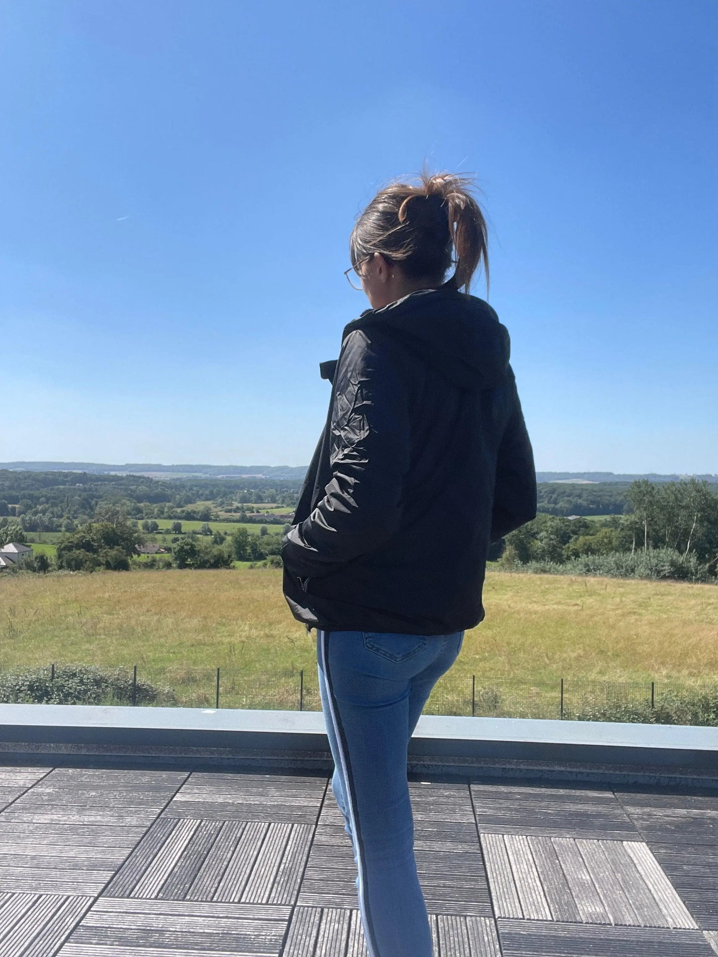 Woman in Laura Jo black raincoat and jeans on terrace, overlooking scenic countryside