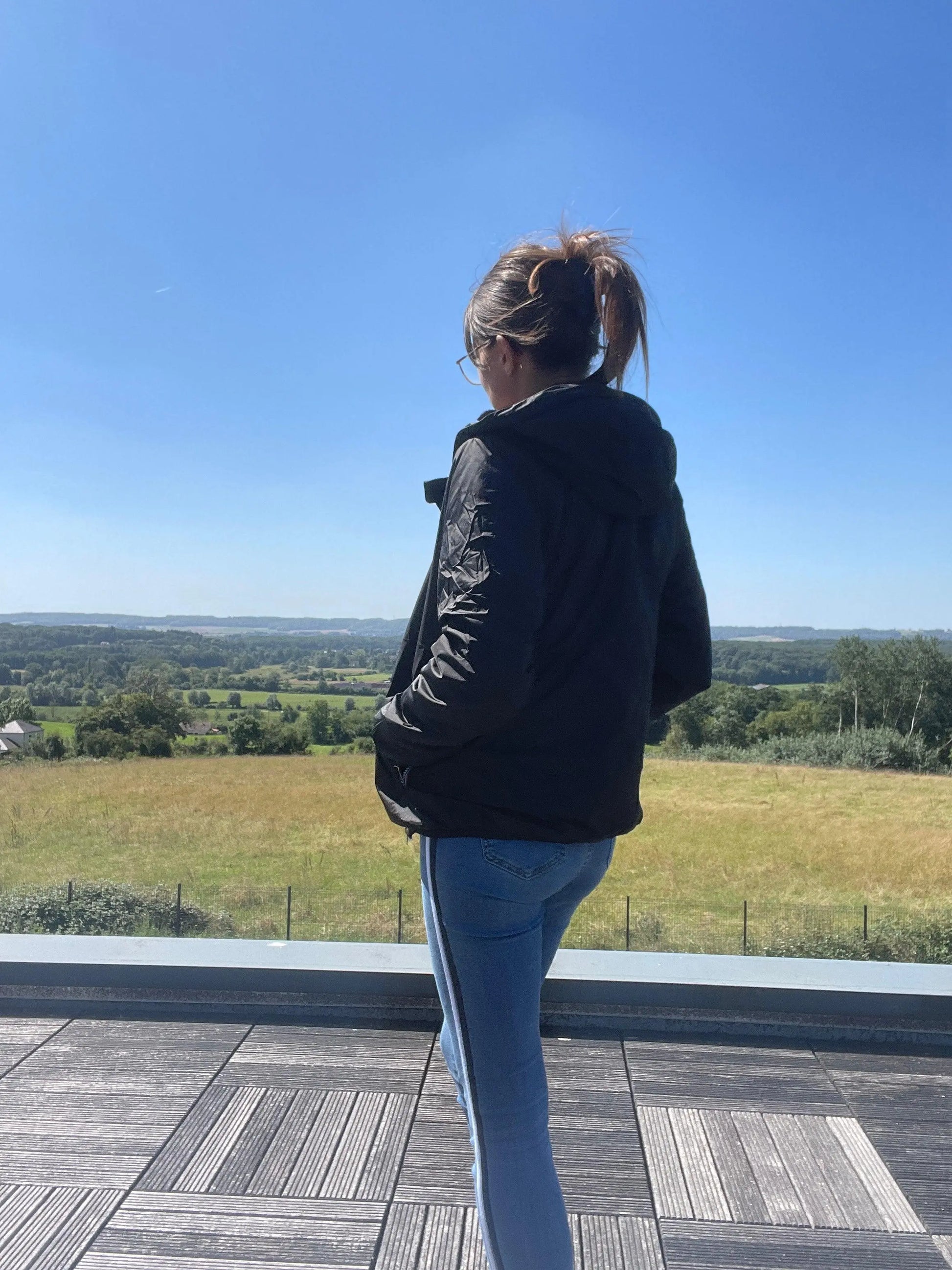 Woman in Laura Jo black raincoat and jeans on terrace, overlooking scenic countryside