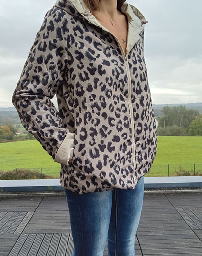 Veste imperméable réversible à motif léopard beige et noir, zip front, col avec capuche, poches, portée par une femme avec jean bleu.