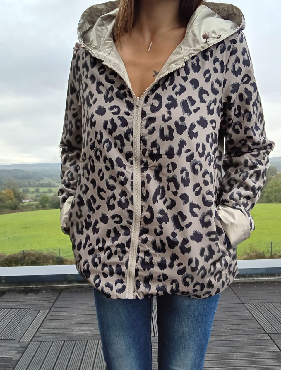 Veste imperméable réversible à motif léopard beige et noir, zip frontal et capuche, porté avec jeans sur fond paysage rural.```