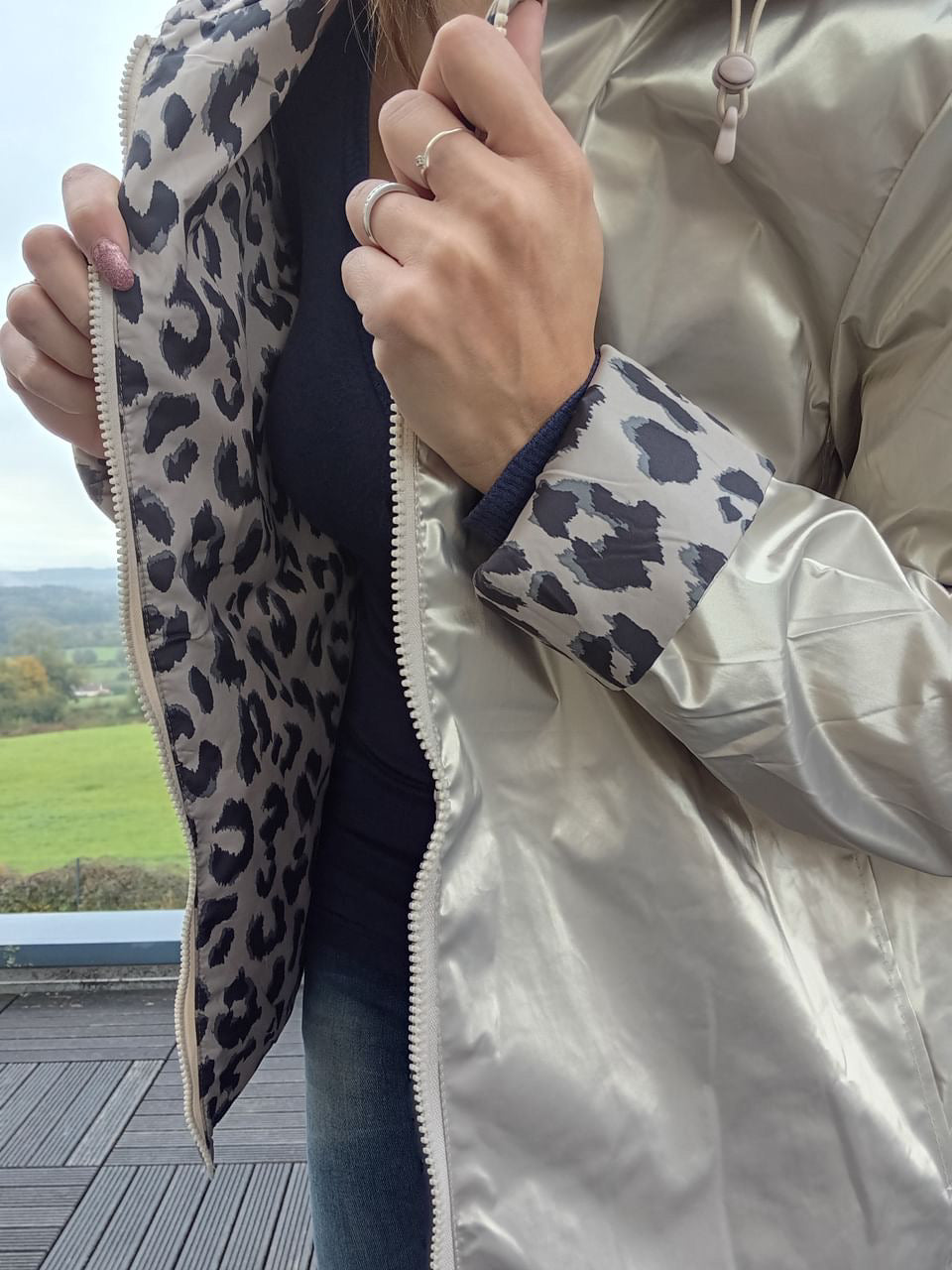 Imperméable réversible clair avec doublure léopard gris à l’intérieur, fermeture éclair visible et cuffs imprimés léopard. Femme montre boutons & doigts avec bague.