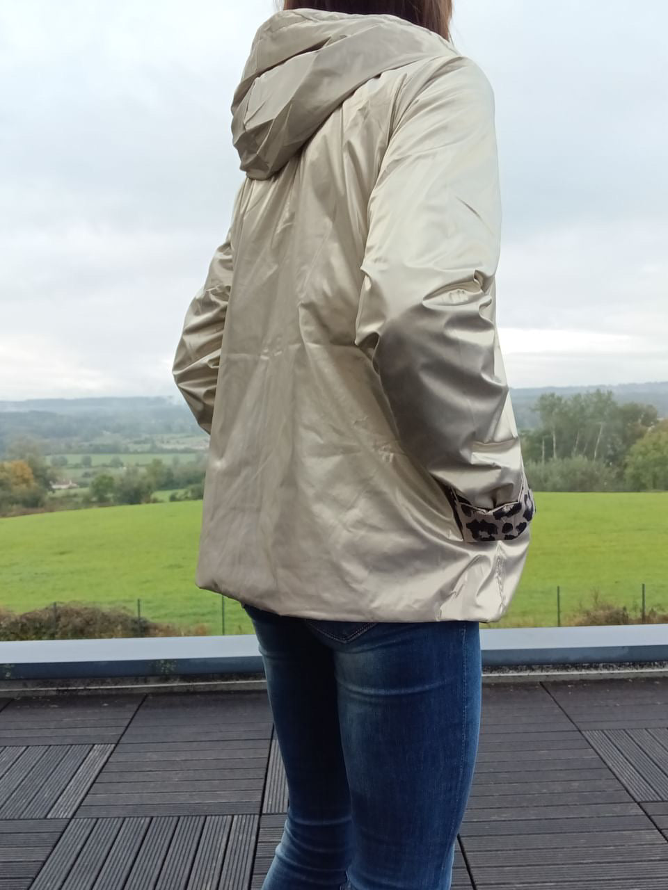 Imperméable réversible court beige, vu de profil, avec capuche, manches longues et poches, porté surbleu jean, paysage champêtre en arrière-plan. de profil