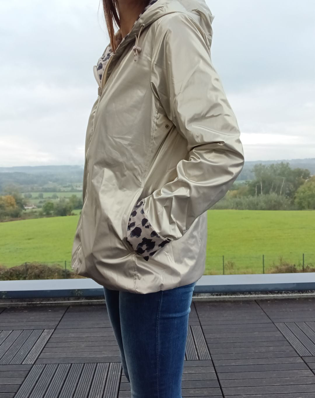 Imperméable réversible couleur beige porté par une femme debout sur deck, poches main, capuche et garnissage mi-saison, paysage champêtre en arrière-plan.