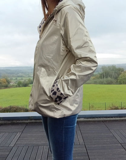 Imperméable réversible couleur beige porté par une femme debout sur deck, poches main, capuche et garnissage mi-saison, paysage champêtre en arrière-plan.