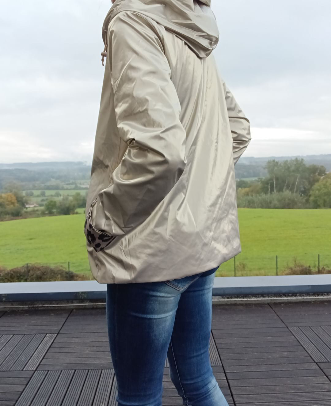 Imperméable réversible court beige, vu de profil, avec capuche, manches longues et poches, porté surbleu jean, paysage champêtre en arrière-plan.