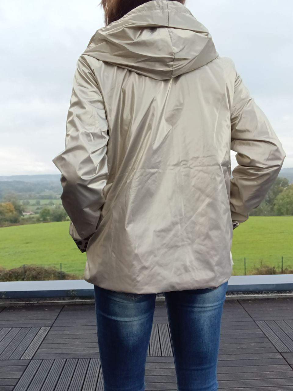 Imperméable réversible beige vu de dos, hoodée, longueur mi-hips, léger garnissage, porté sur jeans, paysage verdoyant en arrière-plan.
