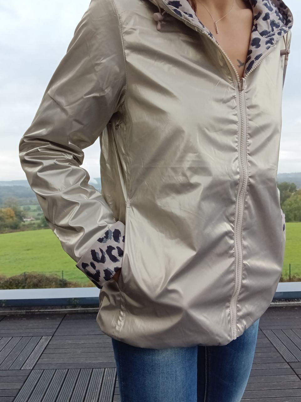 Veste imperméable réversible couleur beige clair, fermeture éclair centrale, motifs léopard à l’intérieur des manches, portée sur fond paysage rural. Altim: imperméable réversible Laura Jo mi-saison.