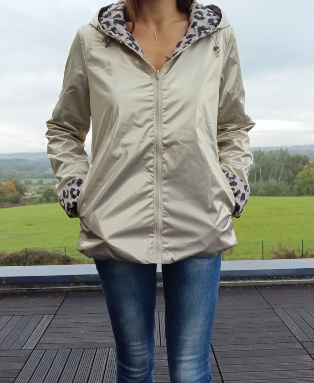 Imperméable réversible beige court avec capuche et doublure Léopard, fermeture éclair frontale, porté en extérieur sur fond paysage rural.