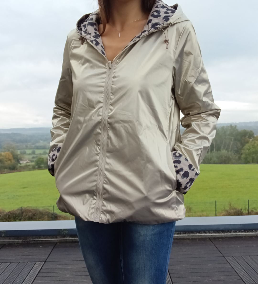 Imperméable réversible court couleur beige avec capuche; doublure à motif léopard visible aux revers et aux poignets, porté en extérieur avec jeans.