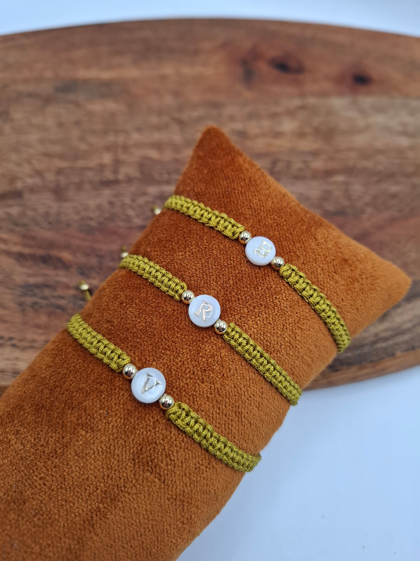 Three green braided bracelets with gold accent beads and lettered white charms on a brown cushion, Le Petit Placard style accessory.