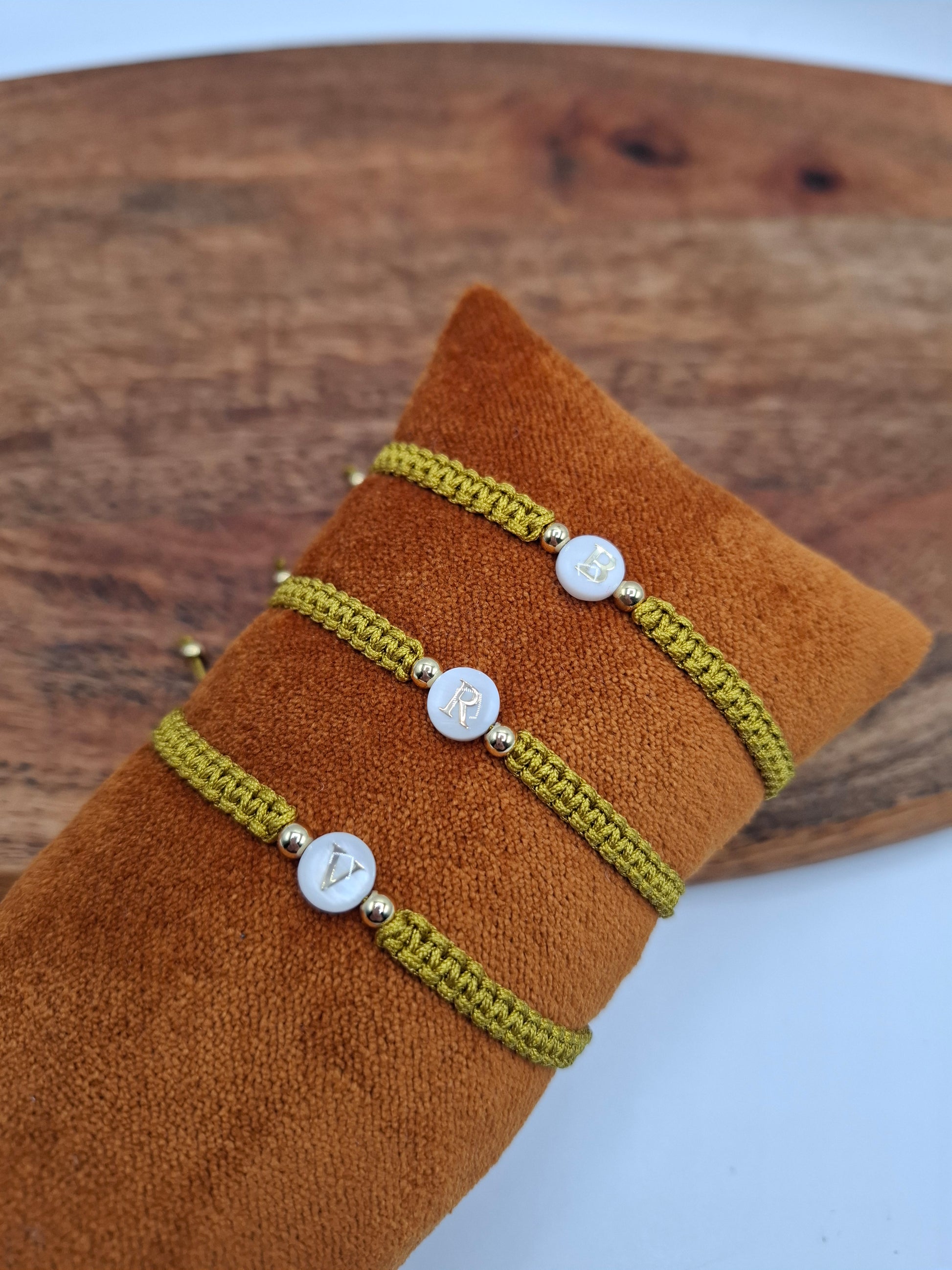 Three green braided bracelets with gold accent beads and lettered white charms on a brown cushion, Le Petit Placard style accessory.