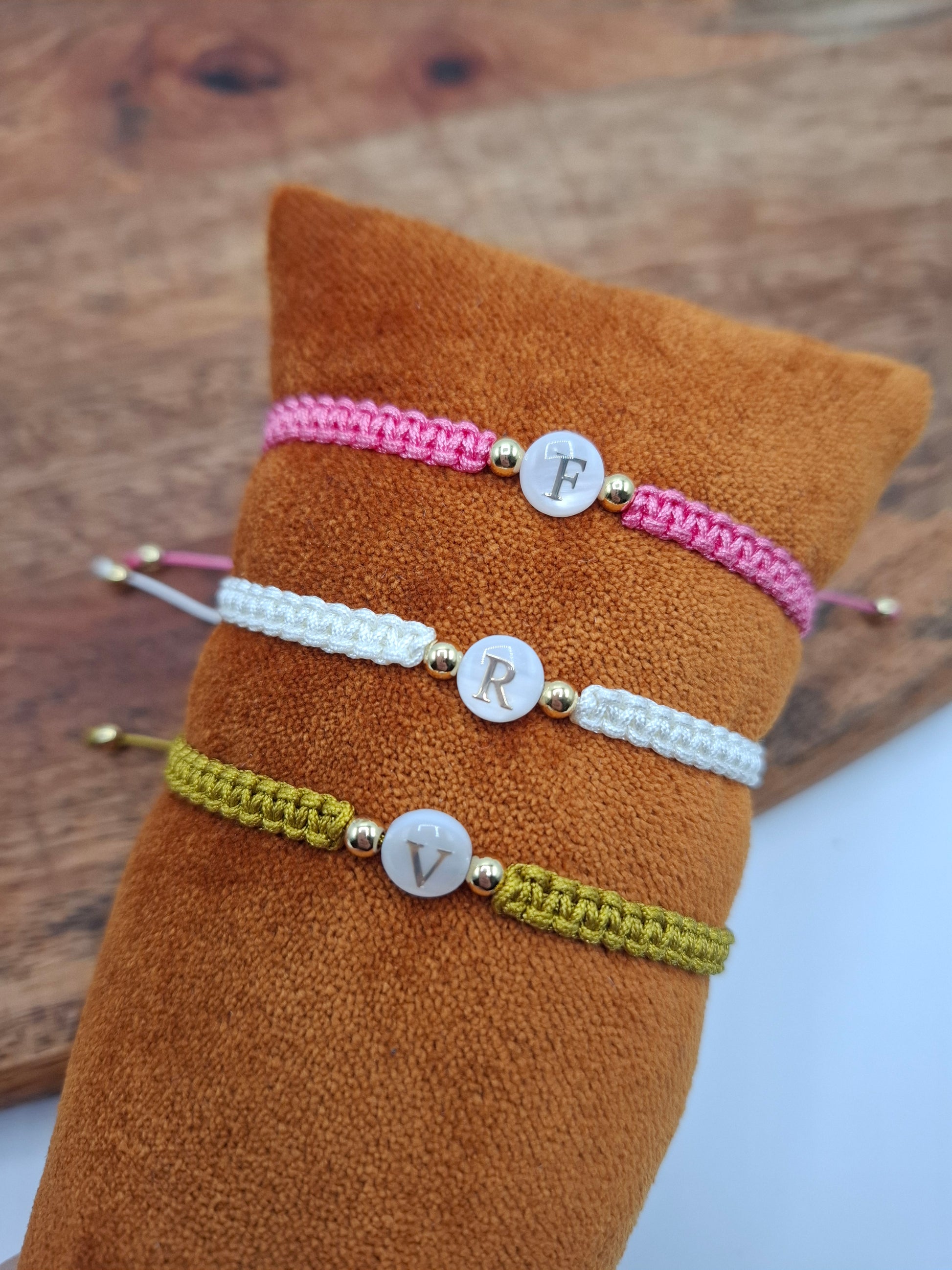 Colorful woven bracelets with gold beads and letter charms displayed on suede cushion, Le Petit Placard.