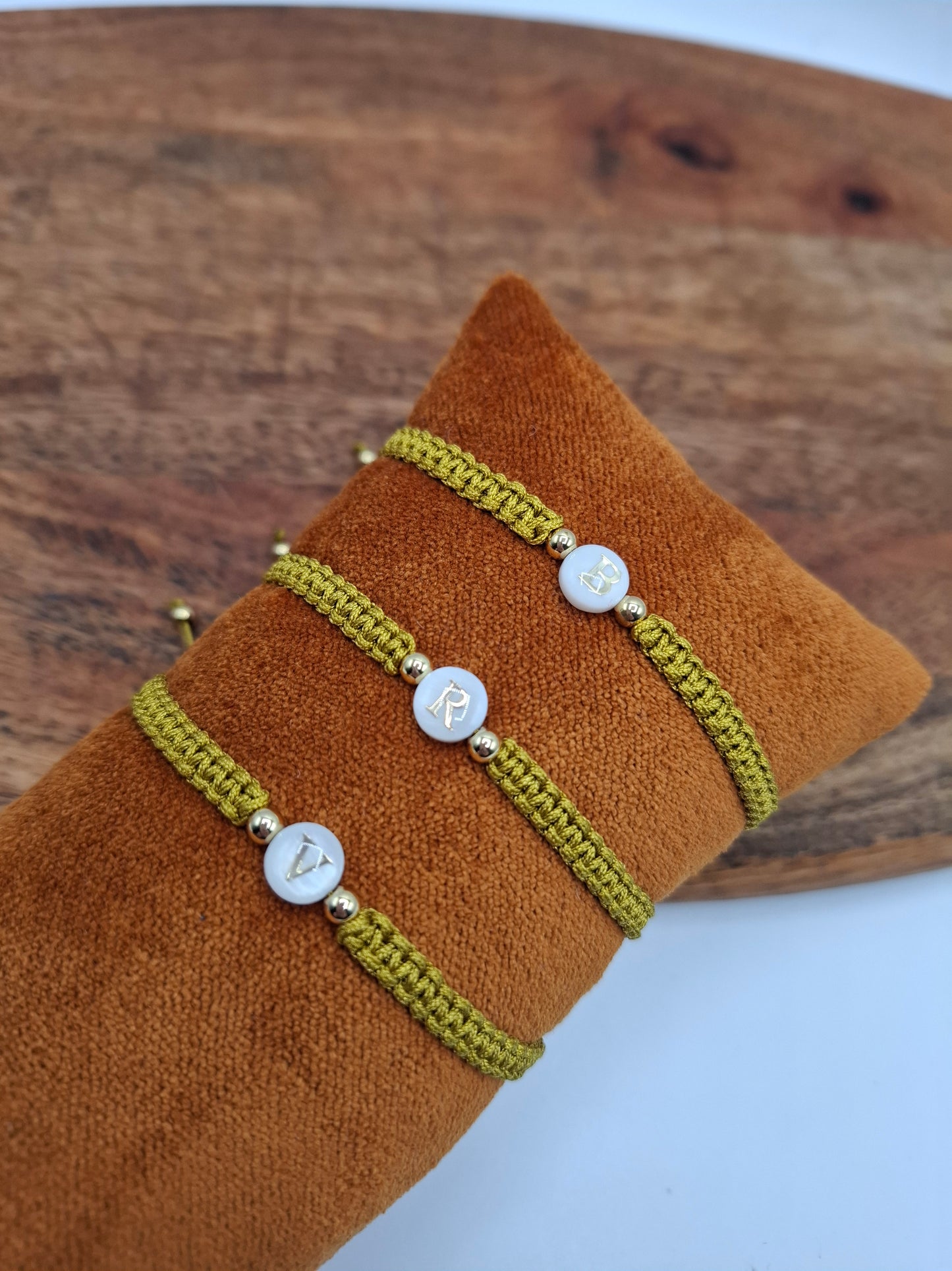 Green woven bracelets with gold accents and letter charms on brown cushion, Le Petit Placard
