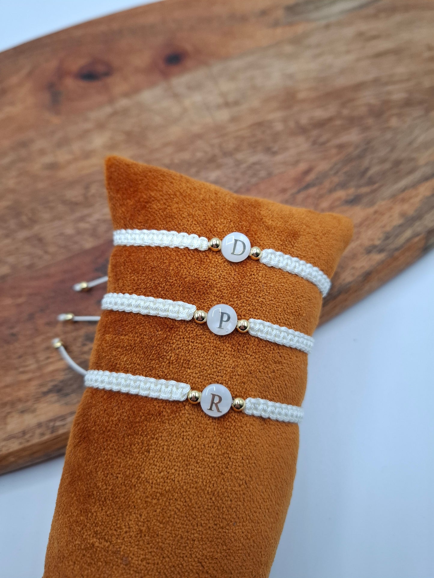 Three white braided initial bracelets with gold beads on a brown cushion, Le Petit Placard accessory