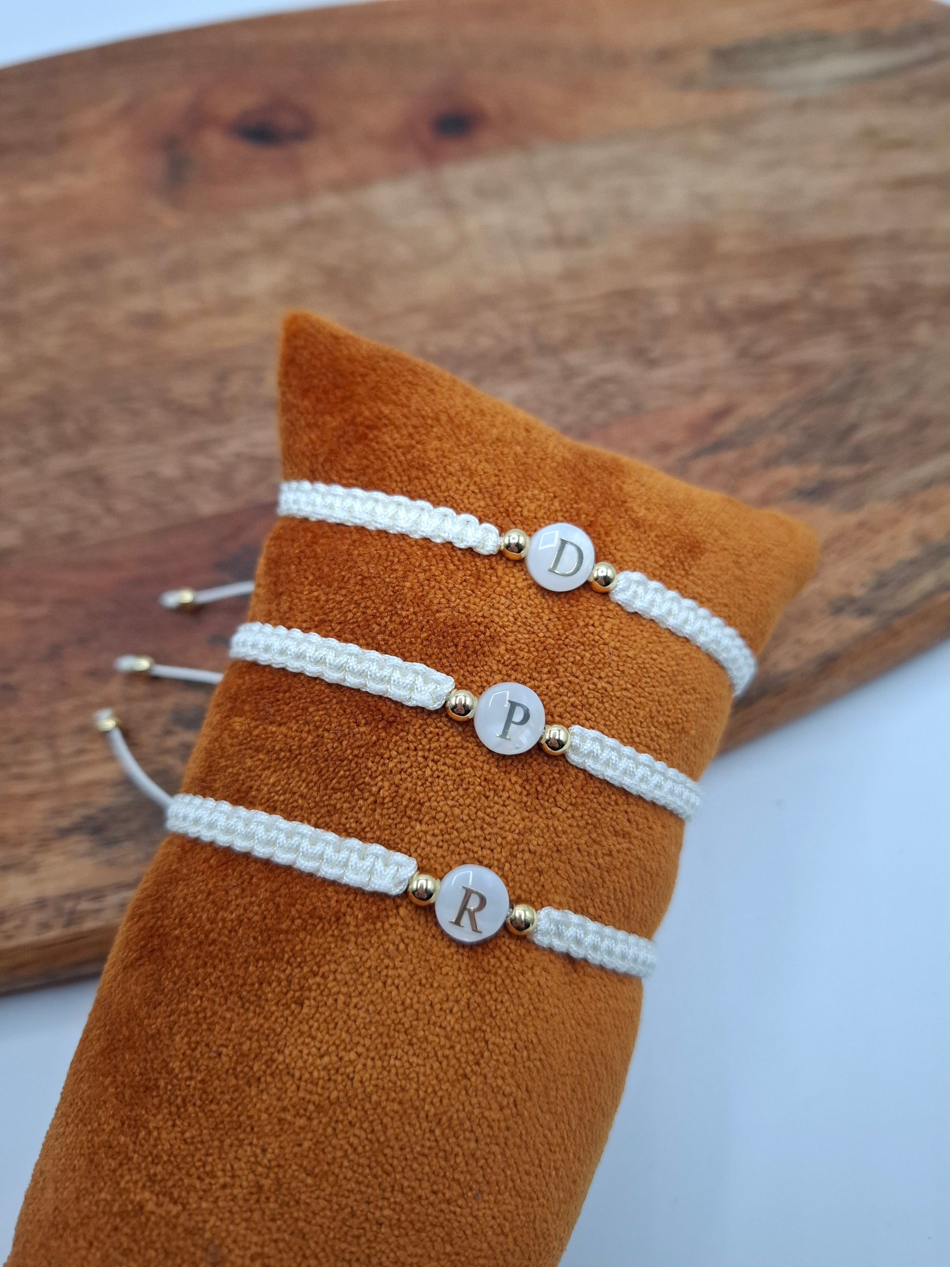 White braided personalized bracelets with gold beads displayed on orange cushion, Le Petit Placard