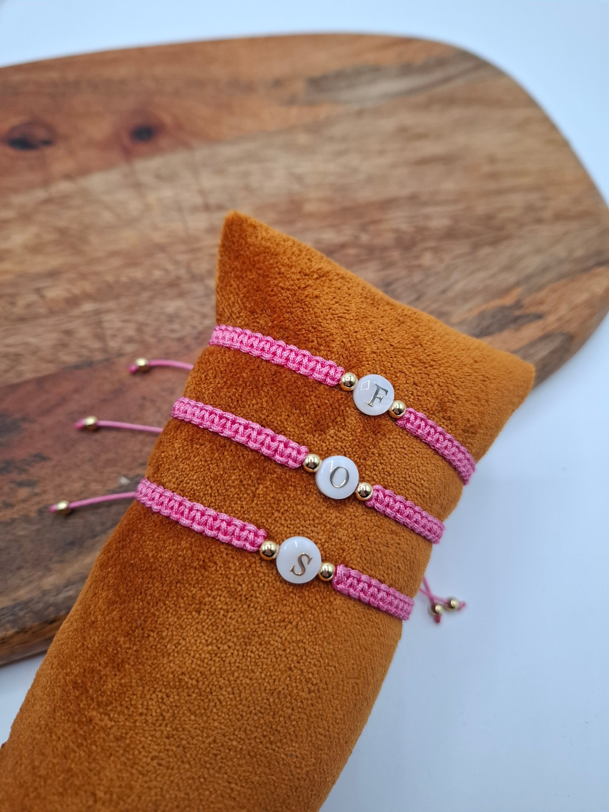 Pink woven bracelets with gold accents and white initial beads, displayed on a suede cushion, Le Petit Placard accessories.