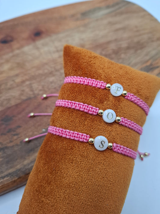 Three pink woven bracelets with gold beads and letter charms displayed on a velvet cushion.