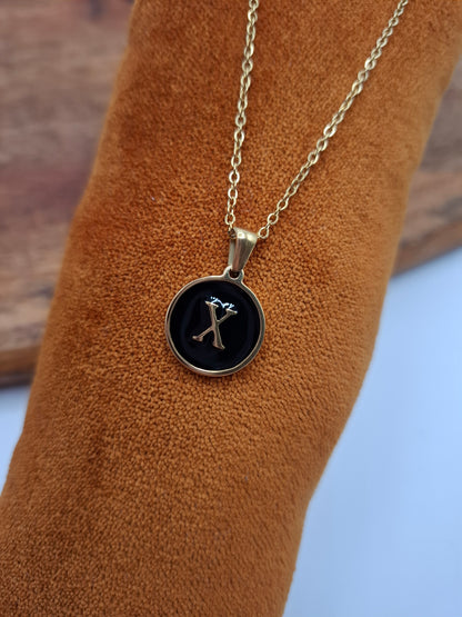 Gold pendant necklace with black round charm and letter X, displayed on brown fabric