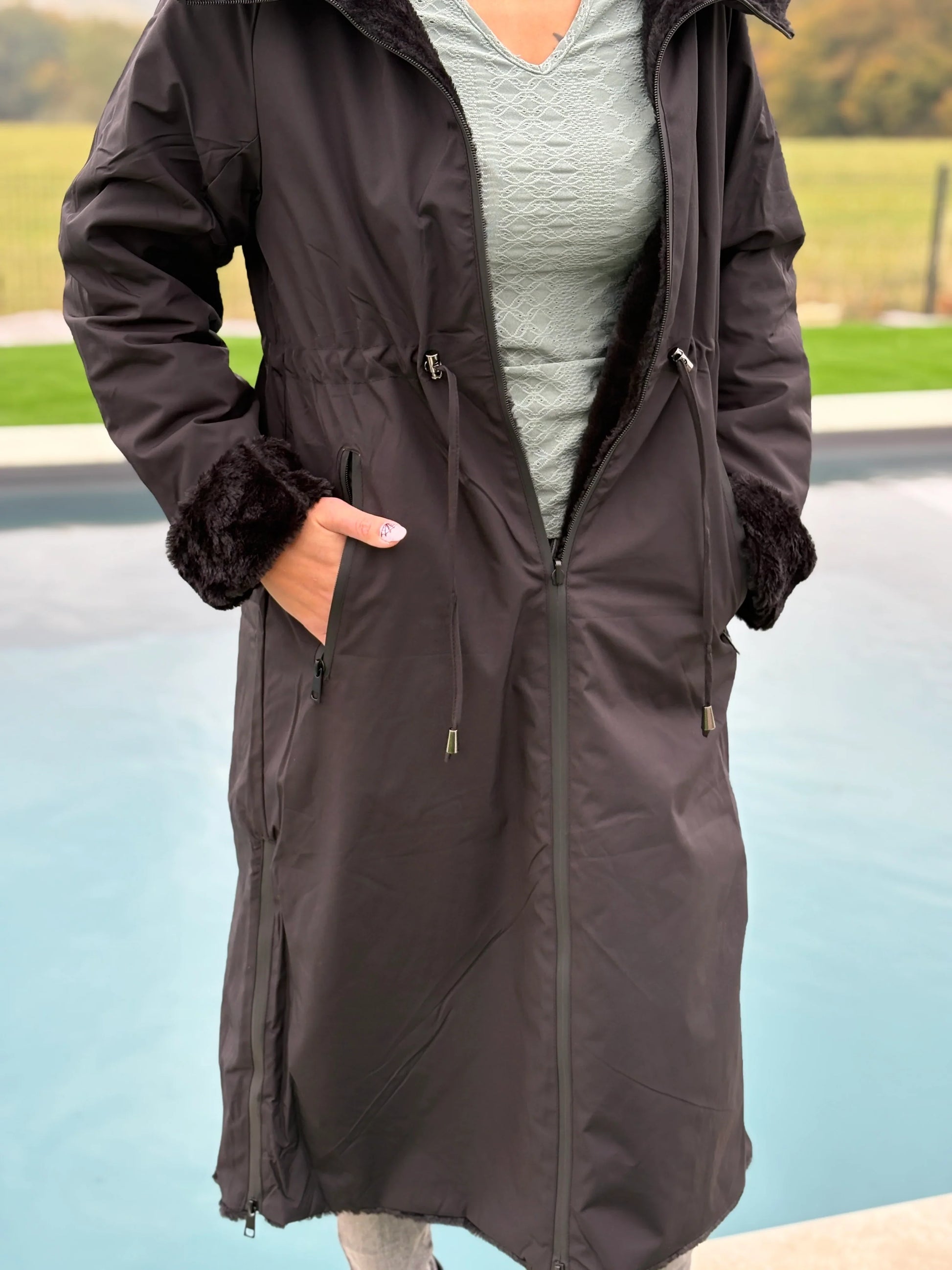 Manteau long réversible noir en fausse fourrure avec capuche et coupe ajustée, porté par une femme.