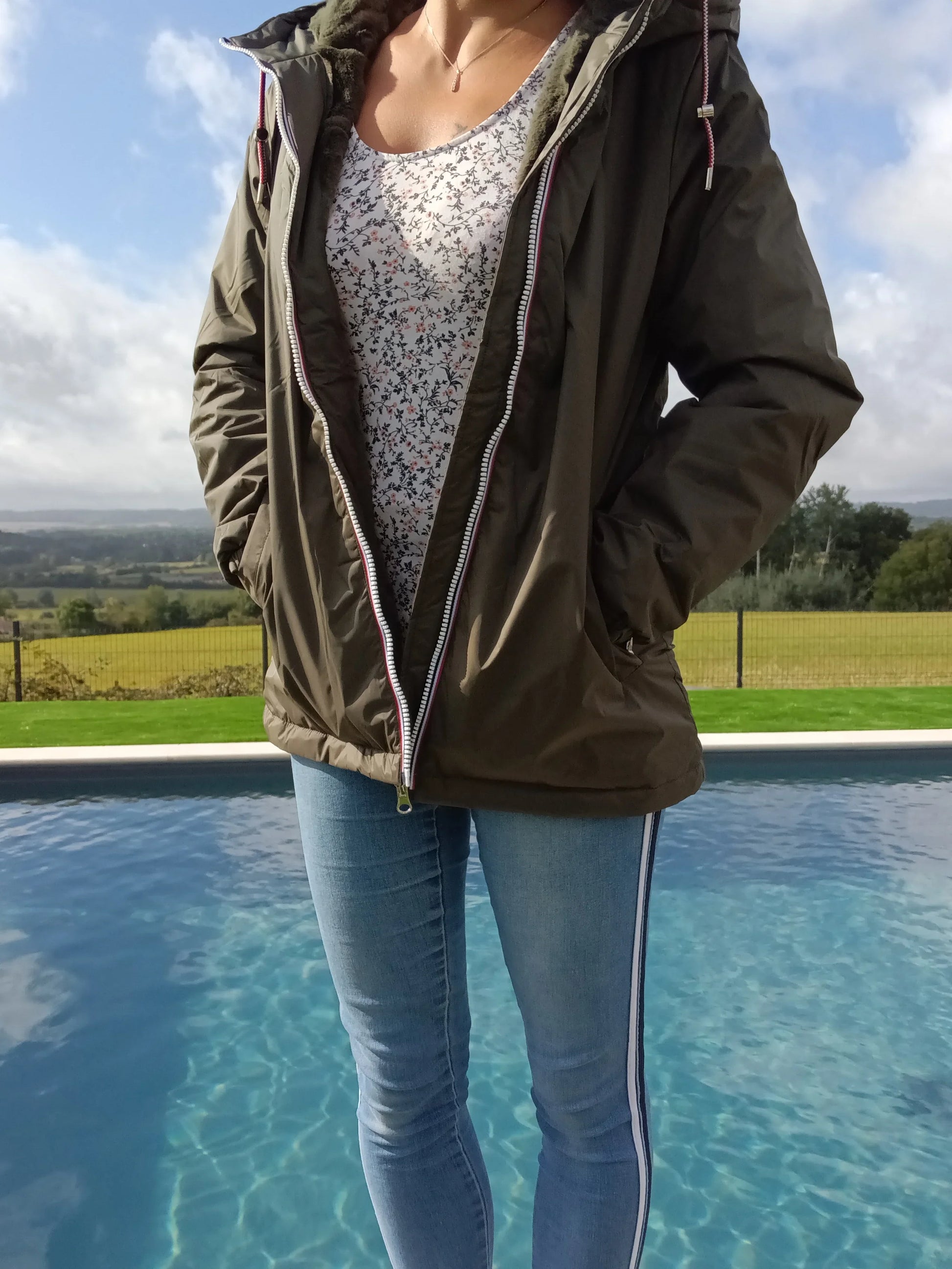 Woman in olive Laura Jo raincoat, floral top, and jeans by pool, outdoor fashion.