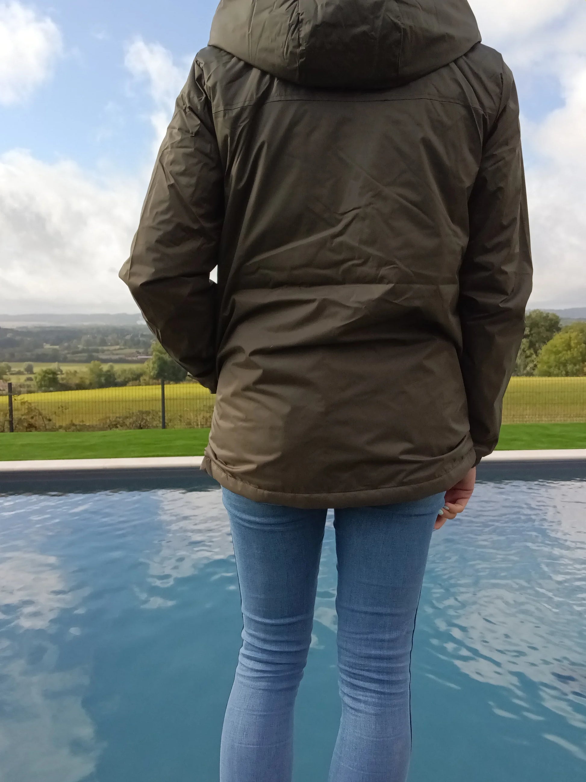Person in green Laura Jo raincoat and jeans by poolside, countryside view, Le Petit Placard