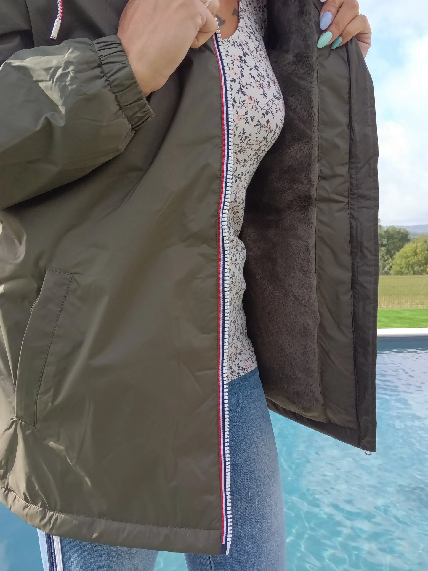 Woman in green Laura Jo raincoat with furry lining, floral top, and jeans by a pool outdoors