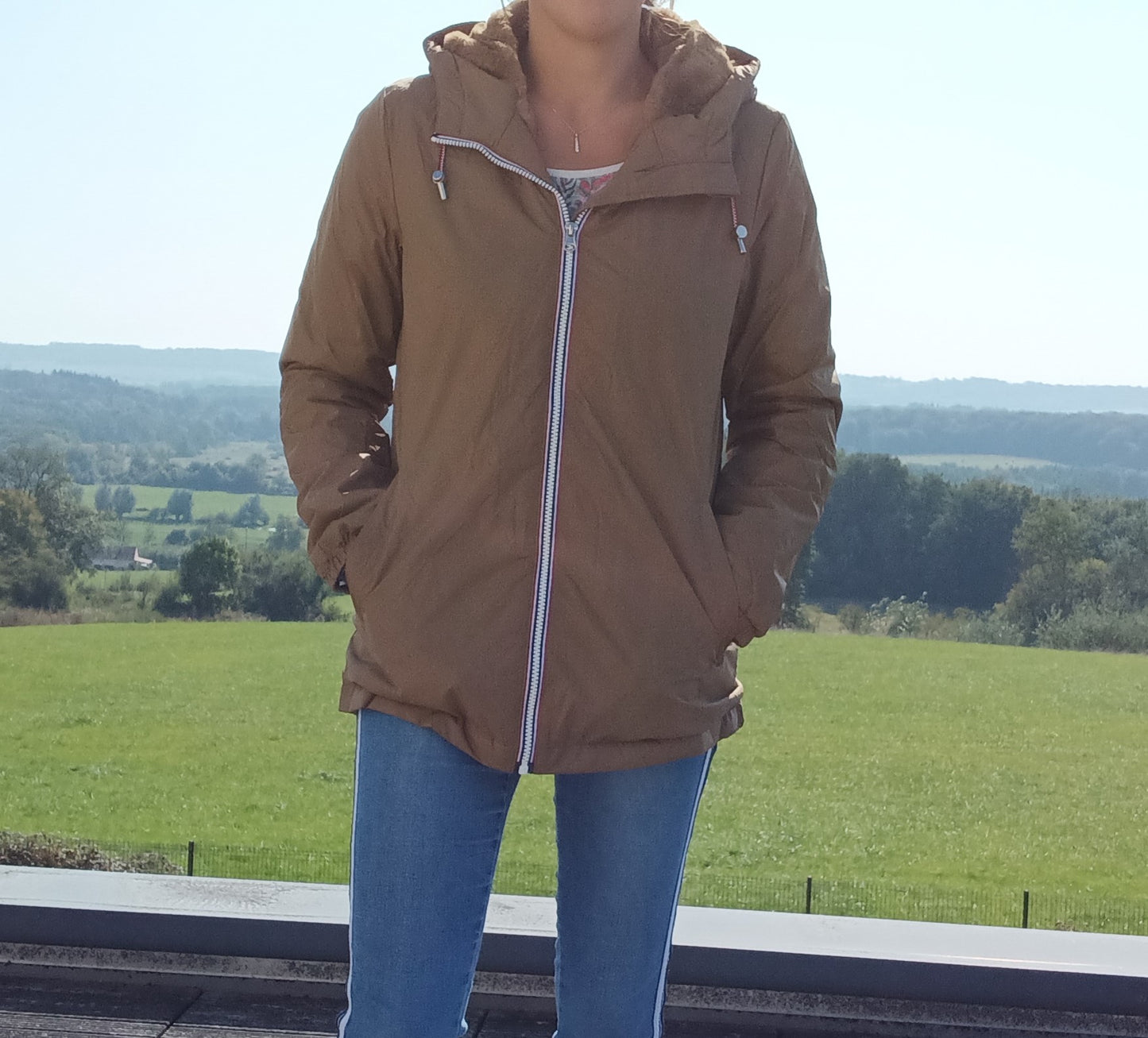 Woman wearing camel Laura Jo raincoat with hood, blue jeans, outdoors with green field