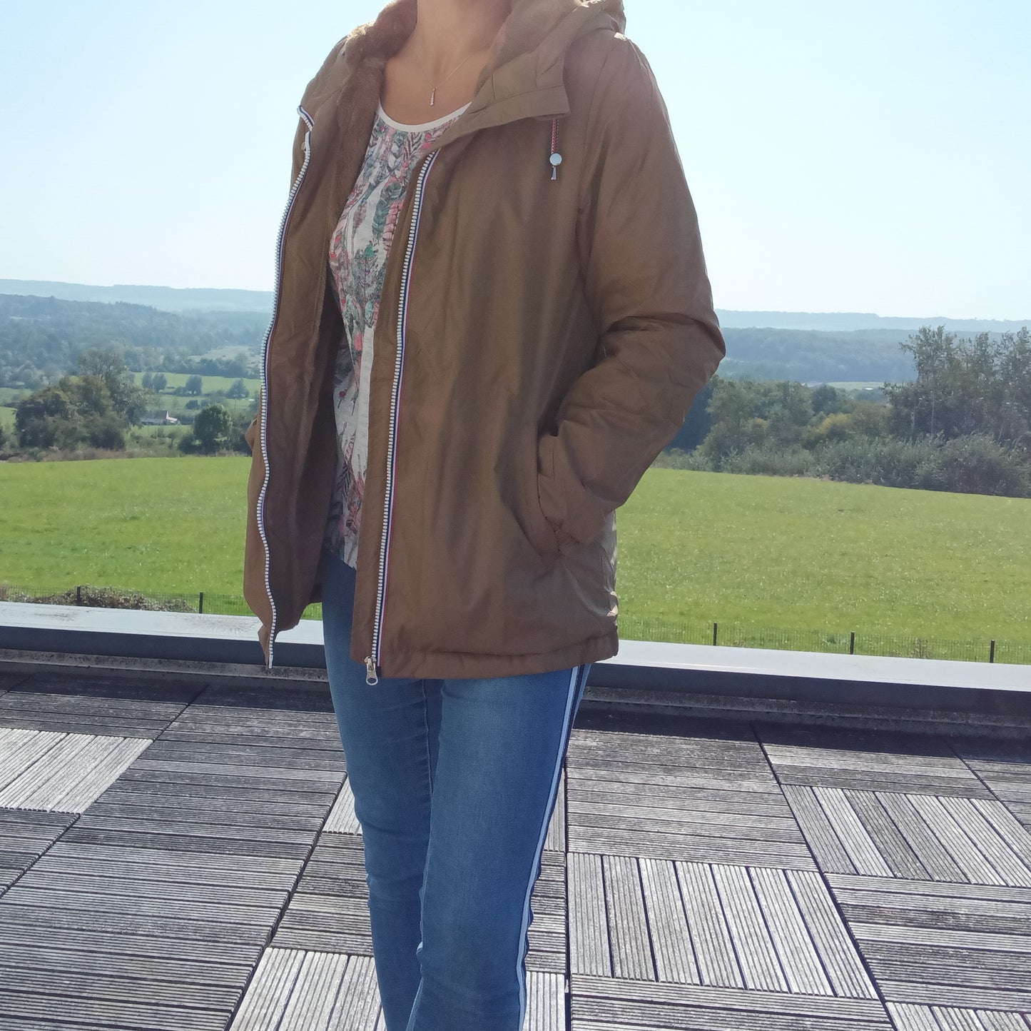 Woman in a camel Laura Jo raincoat and jeans on a terrace with a green countryside view