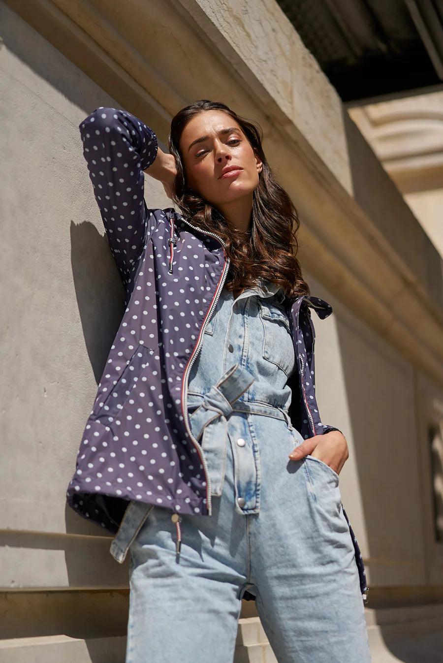 Woman wearing navy polka dot Laura Jo raincoat and denim jumpsuit, leaning on wall outside