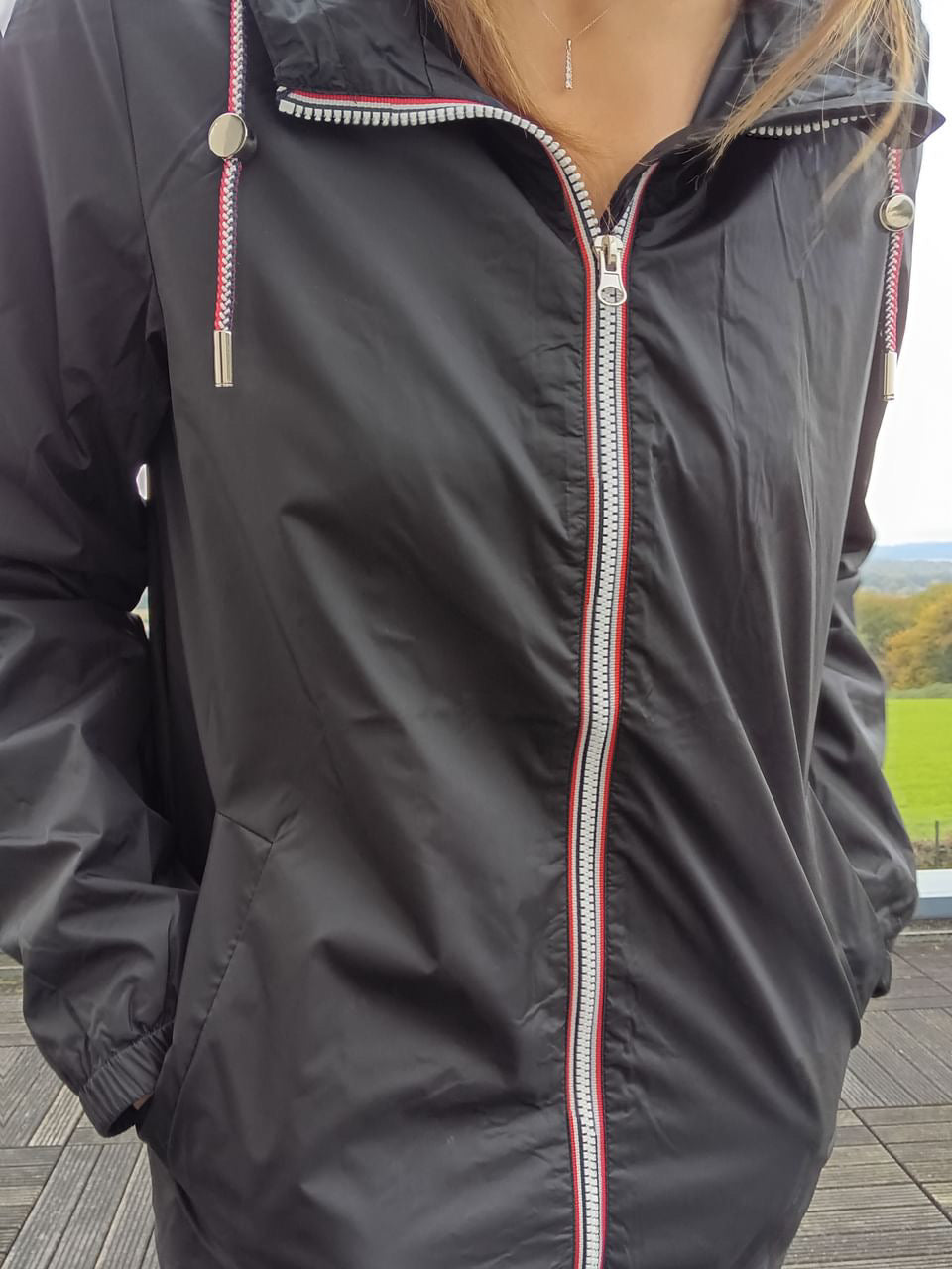 Woman wearing black Laura Jo raincoat with red and white zipper, outdoors on wooden deck